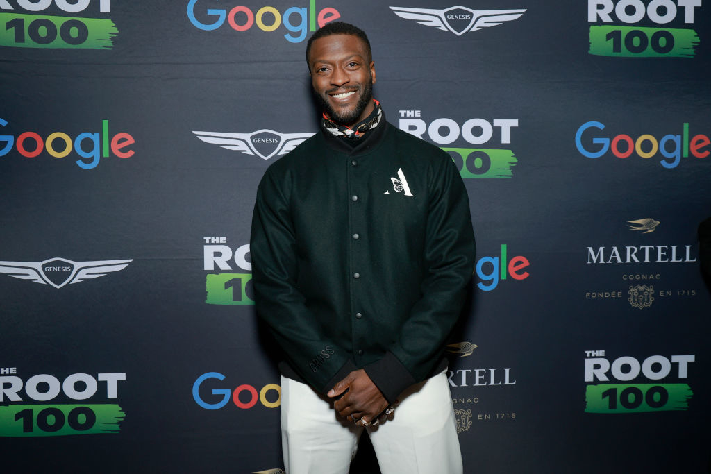 Person standing in front of a backdrop with logos, wearing a dark jacket and light pants, attending a celebrity event