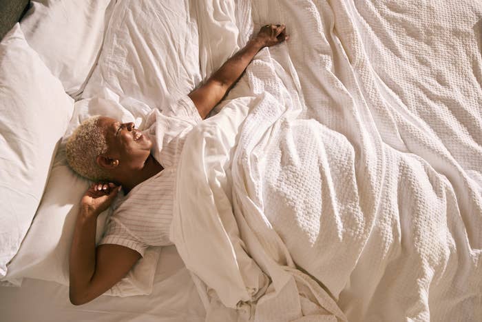 Person relaxing comfortably in bed, lying on their back, smiling slightly. Light is softly illuminating the room