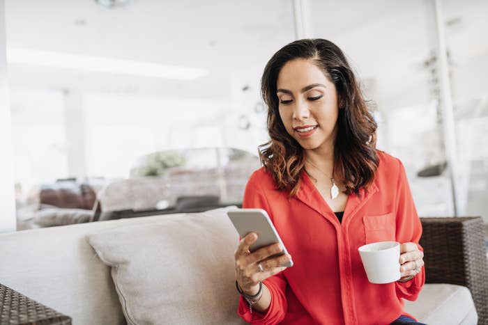 Person sitting on a couch, holding a cup and looking at their phone, wearing a casual, stylish top. Appears relaxed and engaged at home
