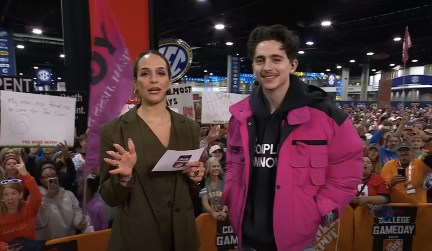 Two people stand indoors at a public event with a cheering crowd in the background. One has a paper, and the other wears a pink jacket over a black hoodie