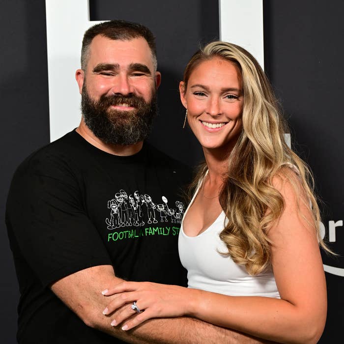 Jason Kelce in casual t-shirt and Kylie Kelce in a white top with satin skirt posing together, smiling on a red carpet