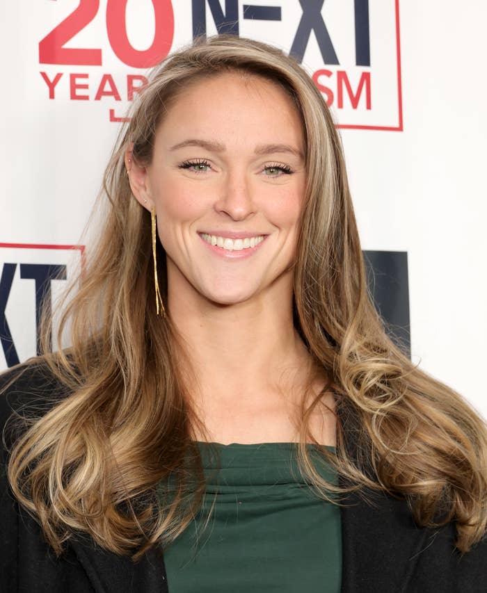 Kylie Kelce smiling at a media event, wearing a black coat over a green top with long hair down