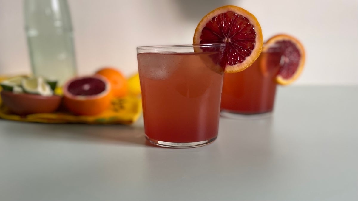 Two glasses of a red-orange drink garnished with a blood orange slice; citrus fruits and a bottle are blurred in the background