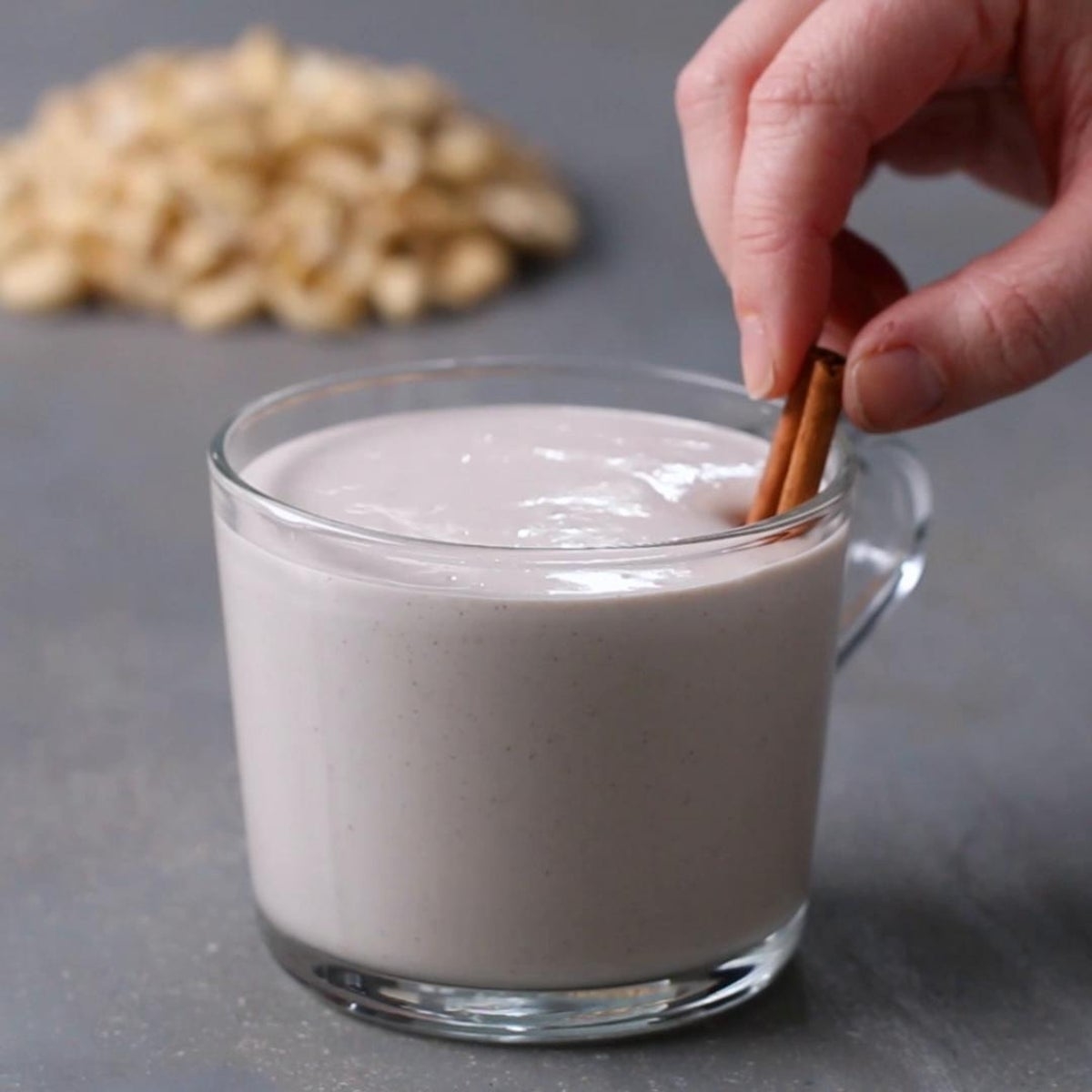 A hand dips a cinnamon stick into a glass mug filled with creamy oatmeal drink. Blurred oats are seen in the background