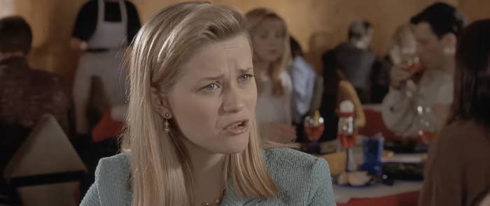 Person with long hair, wearing a textured suit, looks slightly puzzled in a restaurant setting