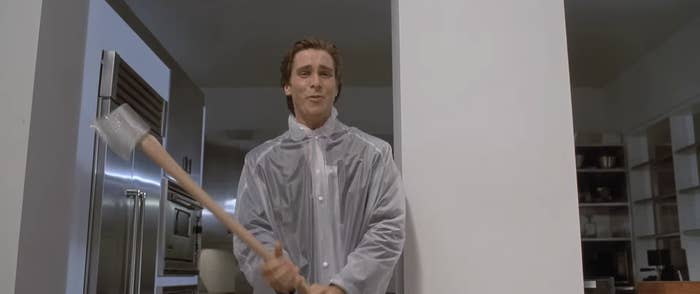 Person in clear raincoat holding axe, standing indoors with a serious expression. Kitchen setting in the background
