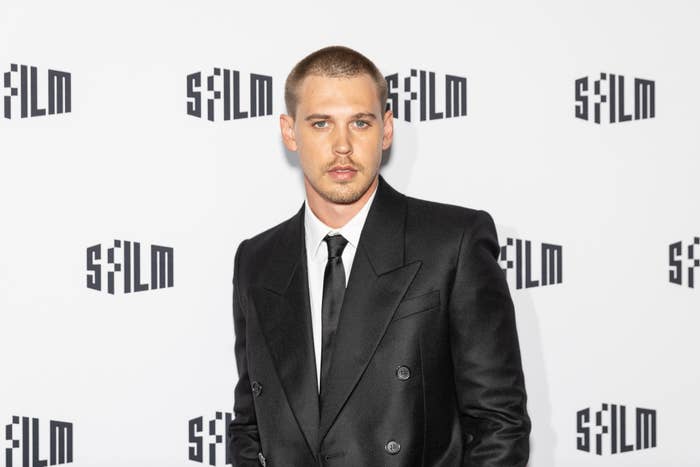Person in formal black suit and tie at a red carpet event in front of a backdrop with 