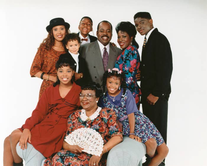 A group of nine people pose together. They are wearing formal and stylish clothing, including hats and floral patterns