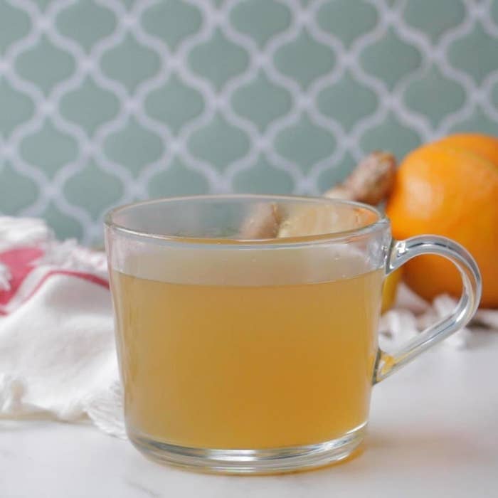 A glass cup of ginger tea on a table, with fresh ginger and oranges in the background