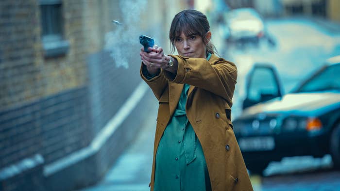 A person in a trench coat points a gun, standing in front of a city street with a parked car