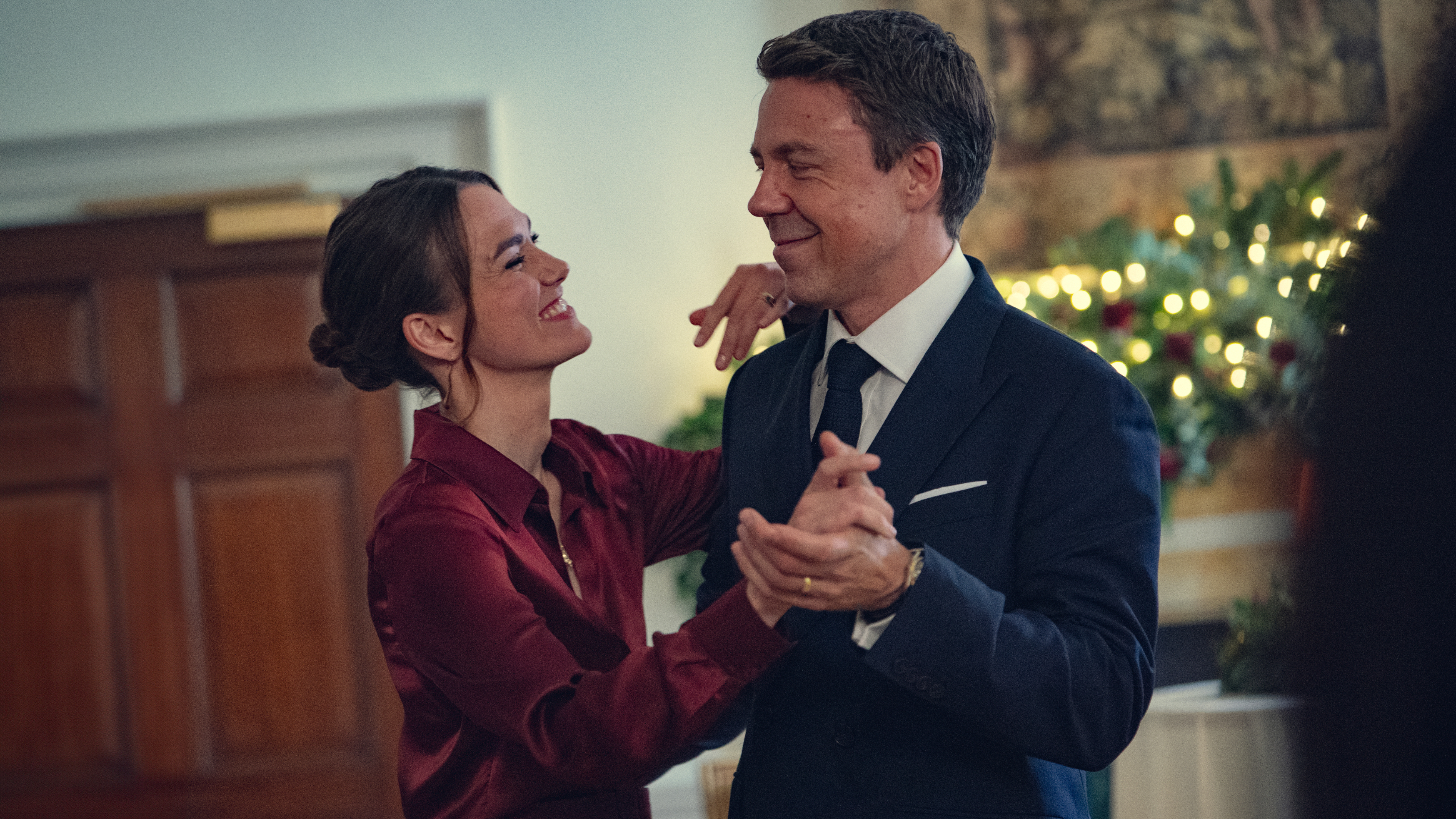 A smiling couple dances in formal attire at an indoor event, with festive decorations in the background