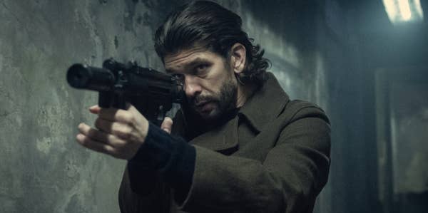 A serious-looking man aims a rifle in a dimly lit, gritty environment, suggesting a scene from an intense TV or movie thriller