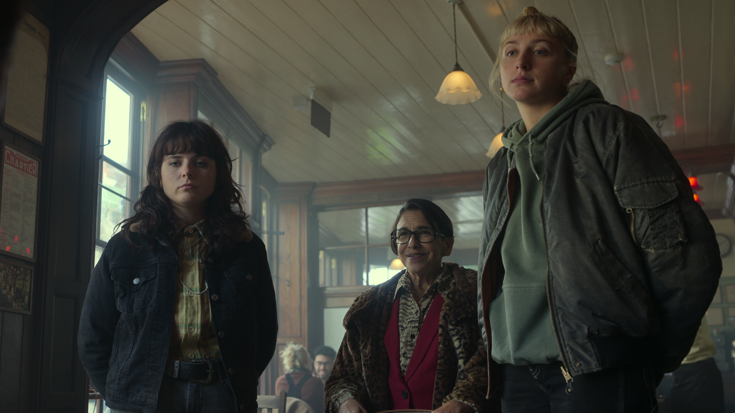 Three people stand in a dimly lit room, wearing casual clothing. The setting appears to be a cozy café