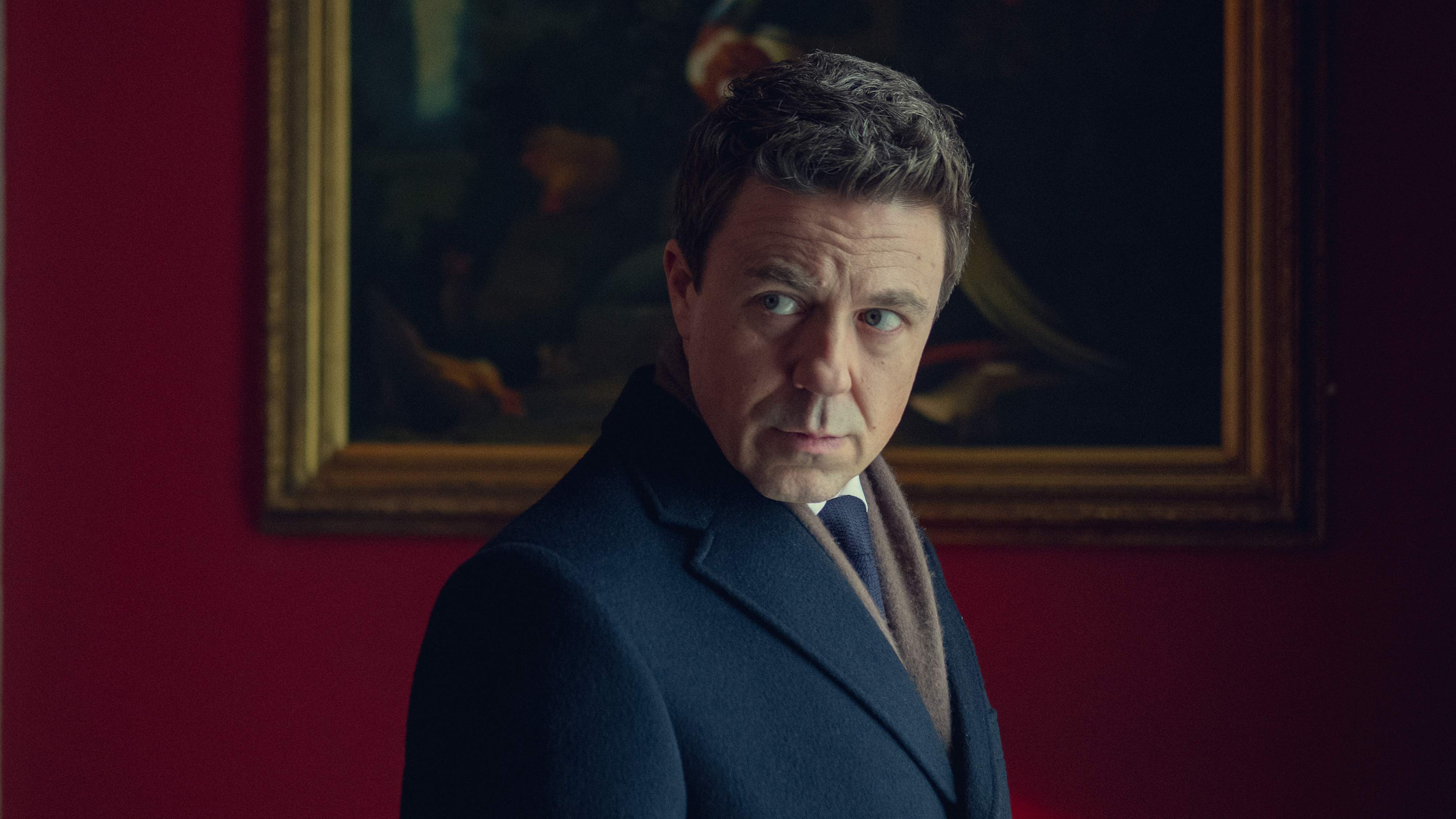 Actor in a formal suit stands in front of a framed painting, gazing thoughtfully to the side