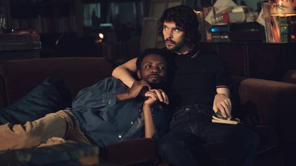 Two people sit closely together on a sofa, one holding a book. The setting suggests a cozy and intimate moment