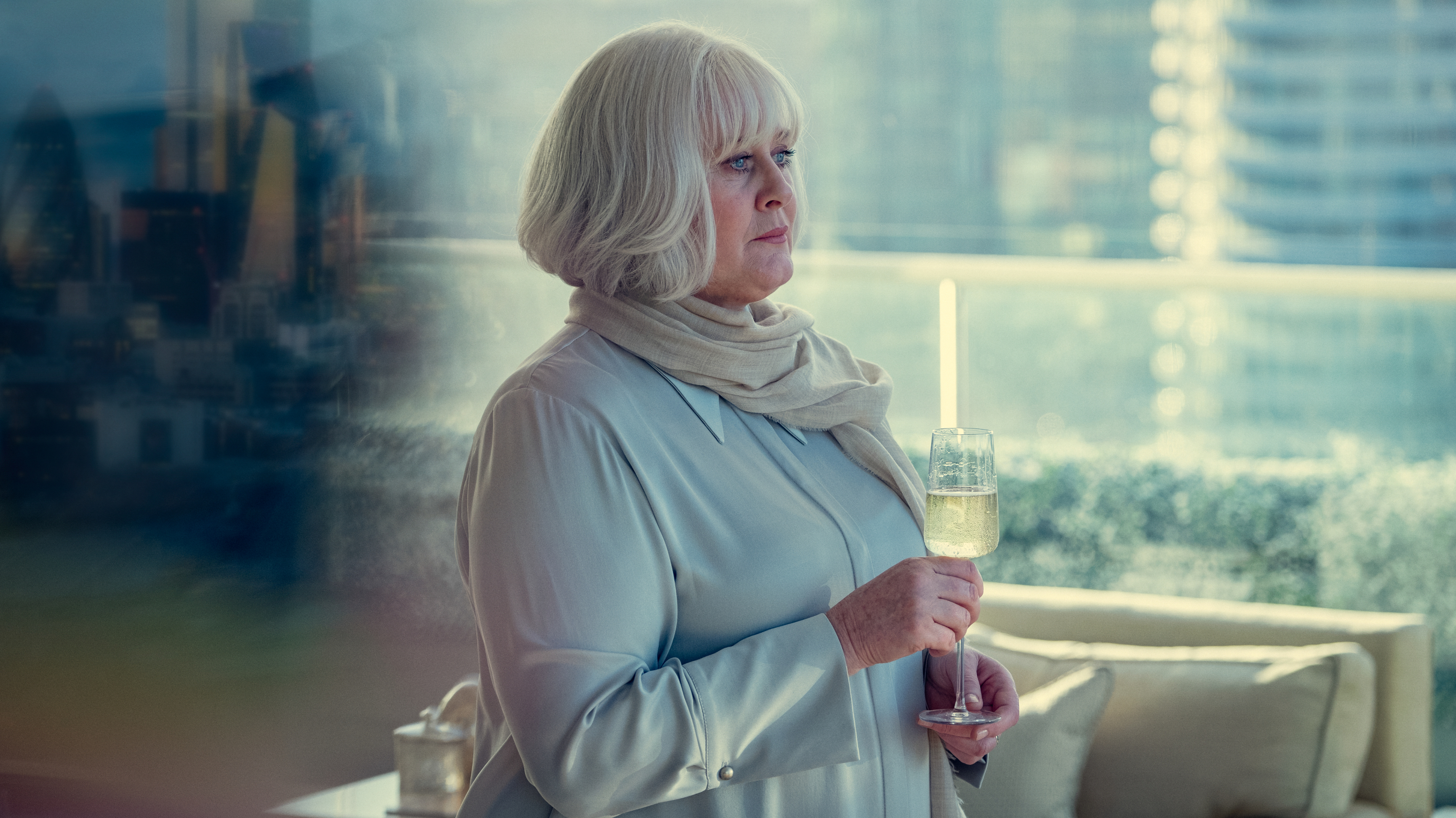 Person with short, light hair, wearing a scarf and holding a glass, stands indoors with a city view in the background