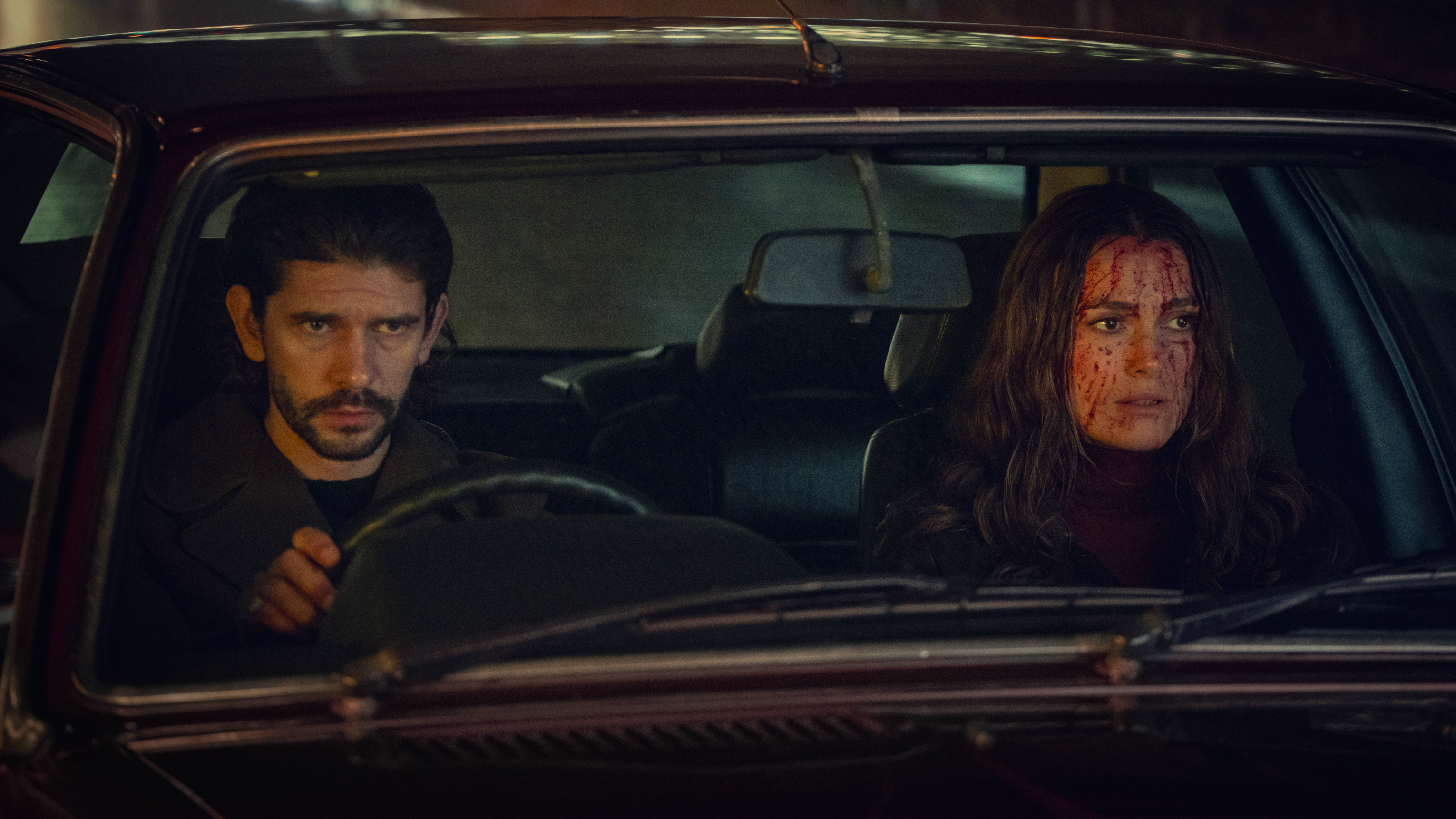 Two people sit in a car at night. The driver looks serious, and the passenger has blood on their face. Scene suggests tension or drama