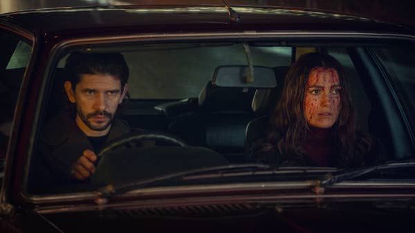 Two people sit in a car at night. The driver looks serious, and the passenger has blood on their face. Scene suggests tension or drama