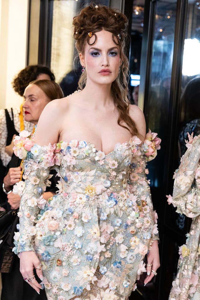 Person in a floral off-shoulder gown with ornate details and an intricate hairstyle stands at an event