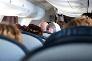 飛行機の客席で帽子をかぶった乗客の後頭部が見える。周囲に他の乗客も座っている。