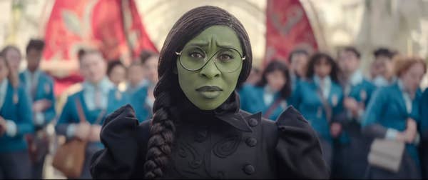 Cynthia Erivo as Elphaba with green skin, wearing glasses and a dark outfit, stands in front of a group dressed in similar school uniforms