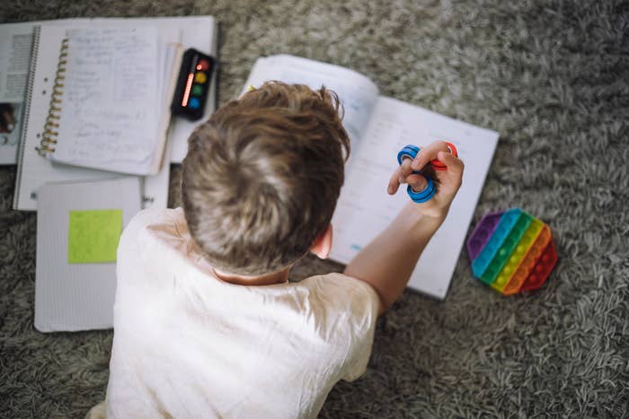 Person sitting on a carpeted floor, holding a fidget spinner, surrounded by open notebooks and a colorful pop-it toy