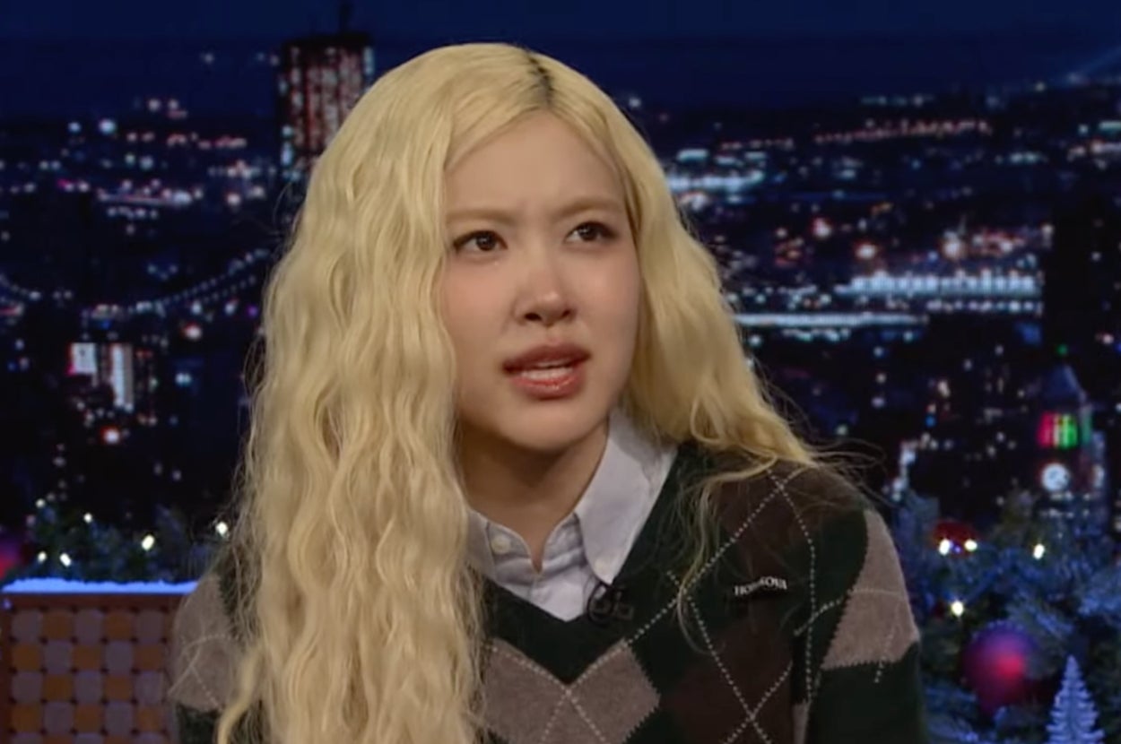 Rosie with long wavy hair in a studio setting wearing a patterned sweater over a collared shirt. City lights are visible in the background