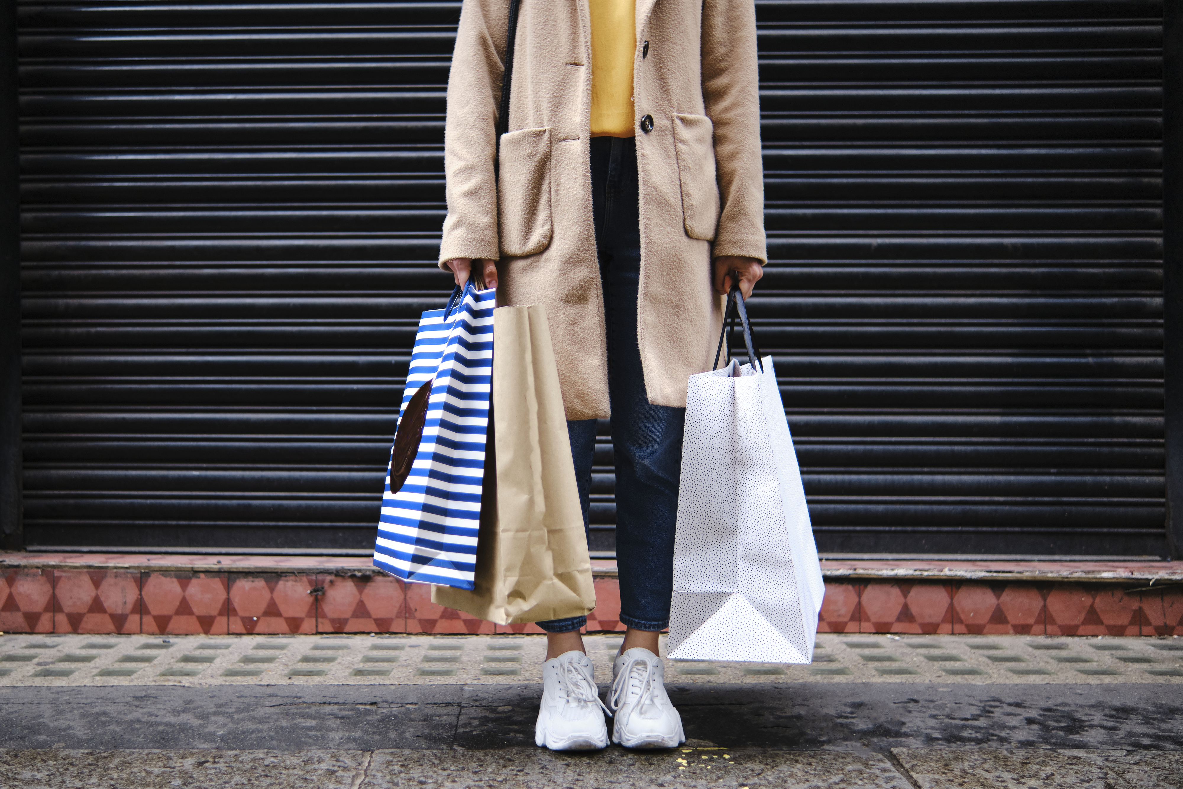 A person holding multiple shopping bags stands in front of a black wall, wearing a long coat, jeans, and sneakers.