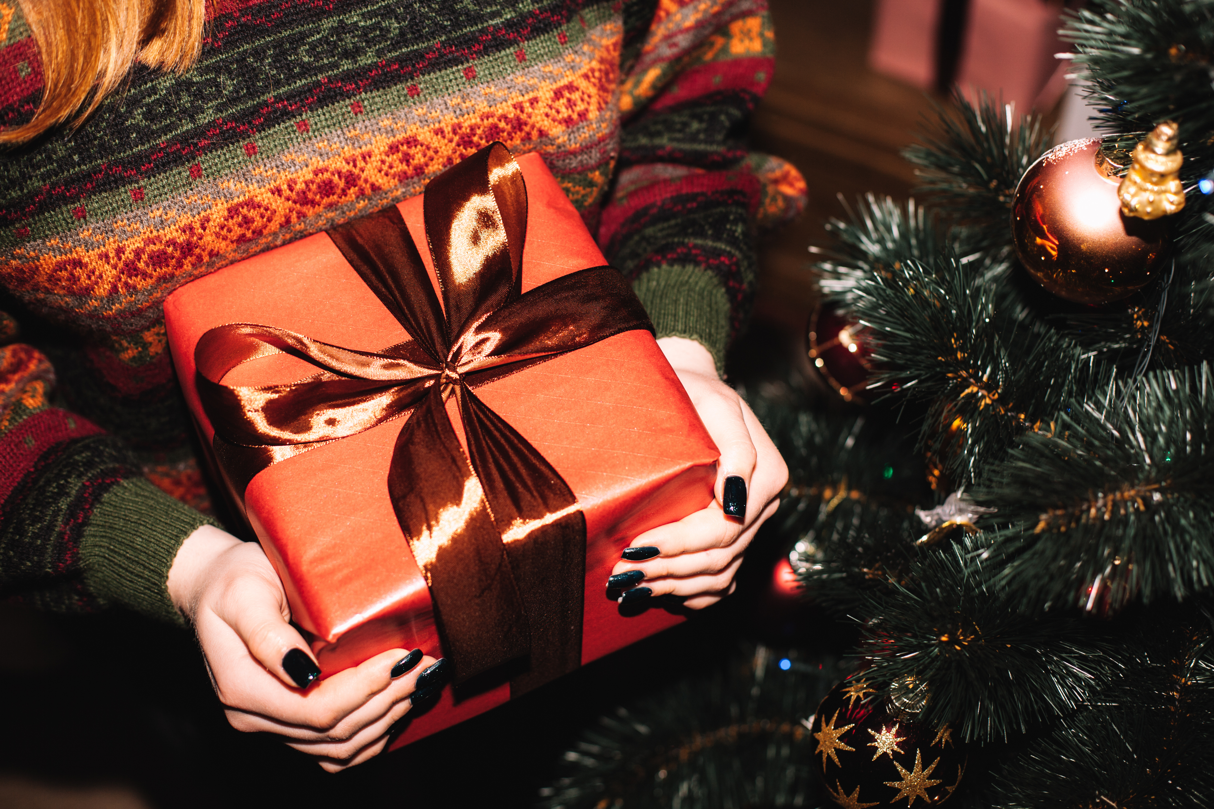 Person holding a gift box with a shiny ribbon, standing next to a decorated Christmas tree.