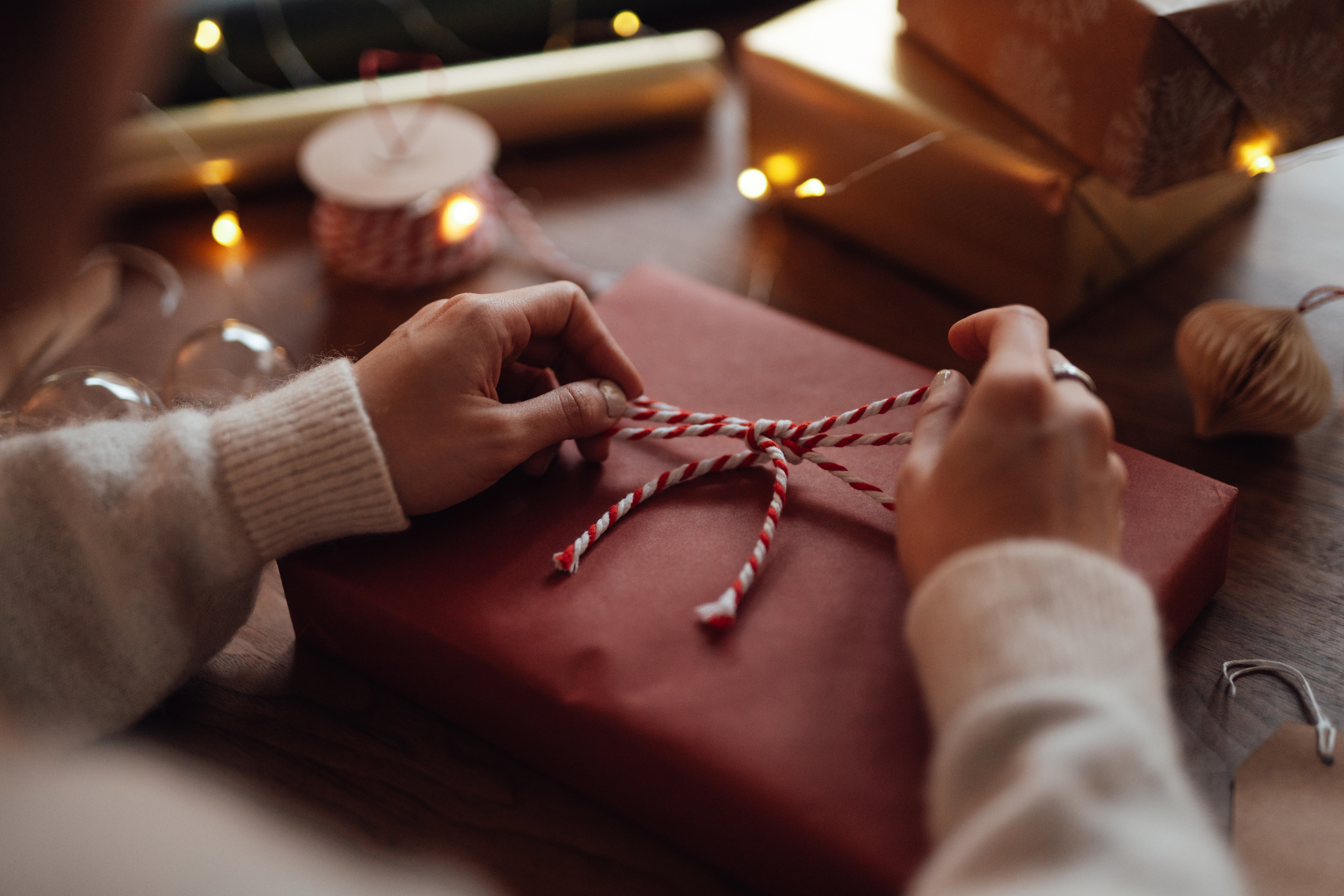 Hands tying a ribbon on a wrapped gift, surrounded by wrapped presents and twinkling lights.