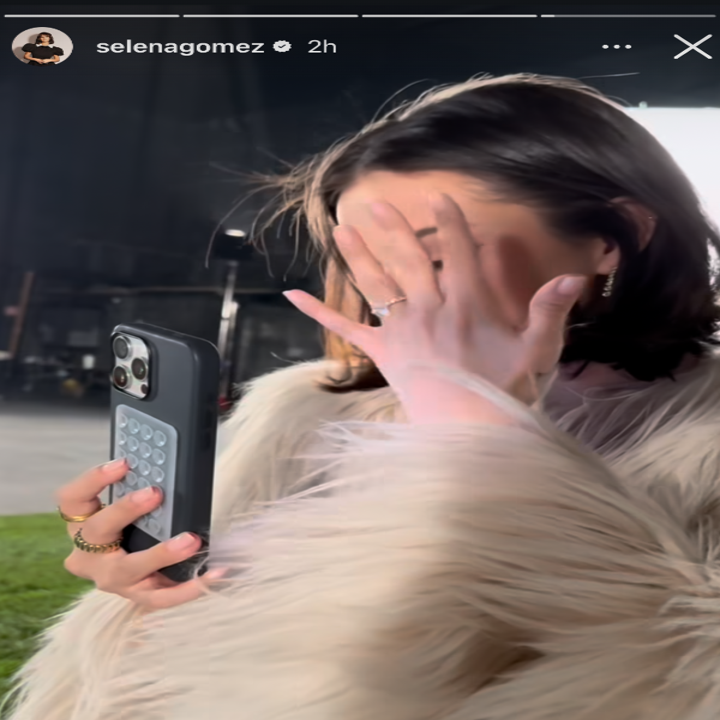 Selena Gomez in a fur coat covers her face with her hand while holding a phone, standing in a dimly lit setting