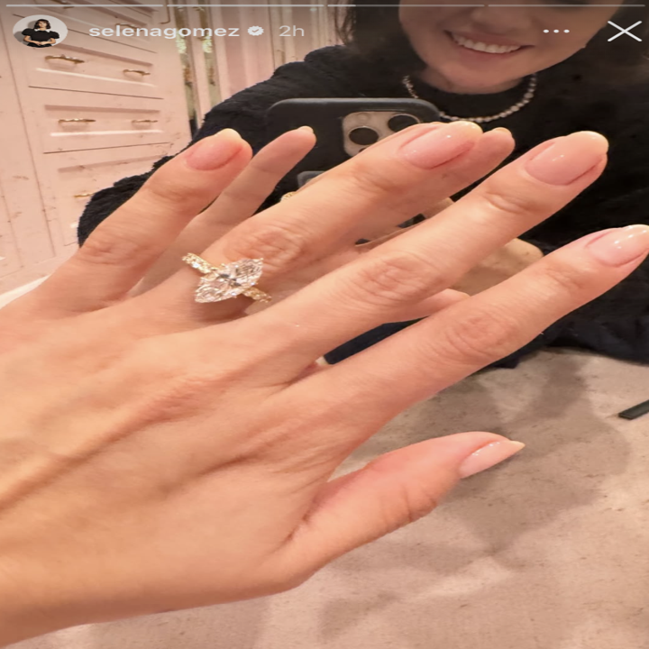 A person displays a large marquise-cut diamond ring, taking a mirror selfie. Only their hand with manicured nails and part of their face are visible