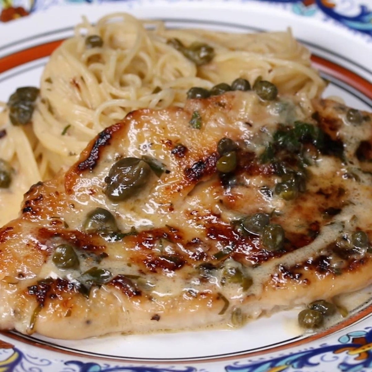 Grilled chicken breast with capers and creamy sauce, served with a side of pasta on a decorative plate