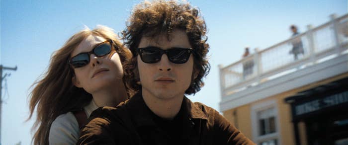 Elle Fanning and Timothée Chalamet wearing sunglasses pose outdoors in a casual setting. One sits on a bike; the other is leaning against them