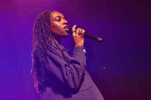 A person performs on stage holding a microphone, wearing a stylish long-sleeve shirt, with braided hair