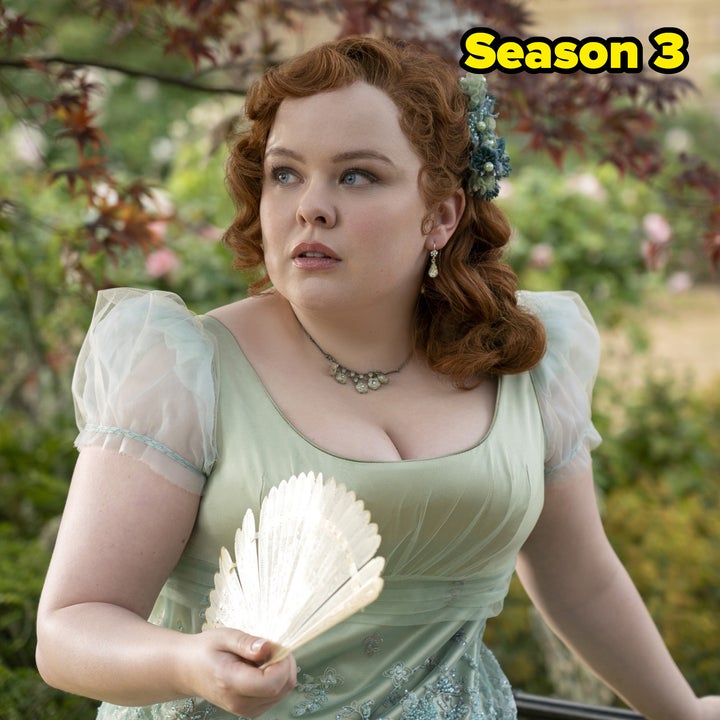 A woman in a Victorian-style dress holds a fan, looking off-camera. She has styled red hair with floral detail