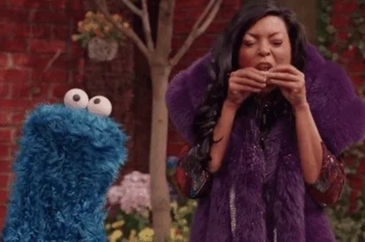 A woman in a fuzzy coat enjoys a cookie next to a blue monster with googly eyes, standing outdoors