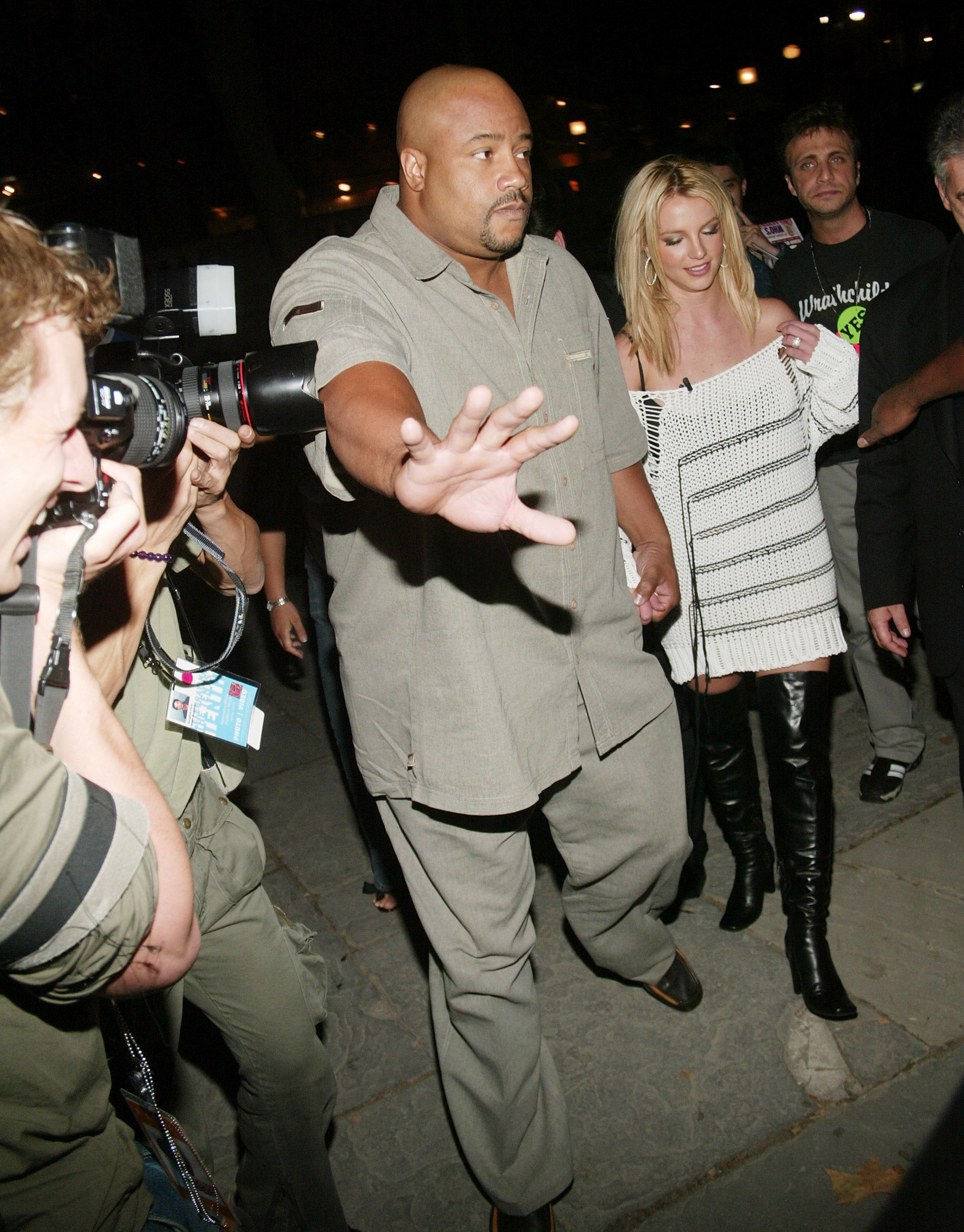 Celebrity escorted through a crowd by a security guard, wearing a knitted dress and knee-high boots, with paparazzi capturing photos