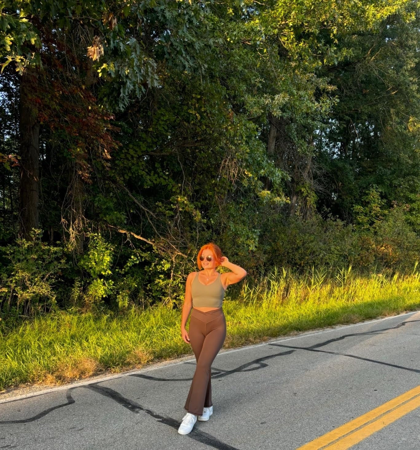 Person walking connected  a paved road, wearing a fitted top, flared pants, and sneakers, surrounded by trees and earthy  greenery