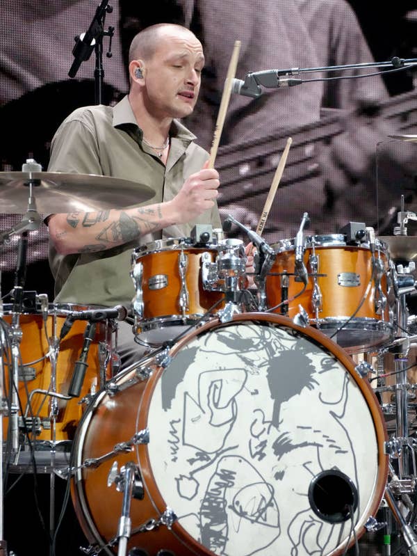George drumming on stage, wearing a short-sleeve shirt. Drum set features artistic doodles on the bass drum