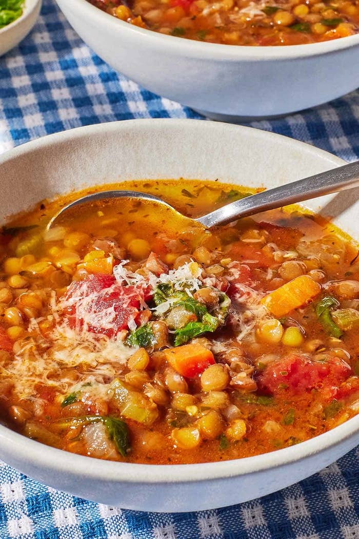 A bowl of vegetable lentil soup with a spoon, topped with grated cheese, on a checkered tablecloth