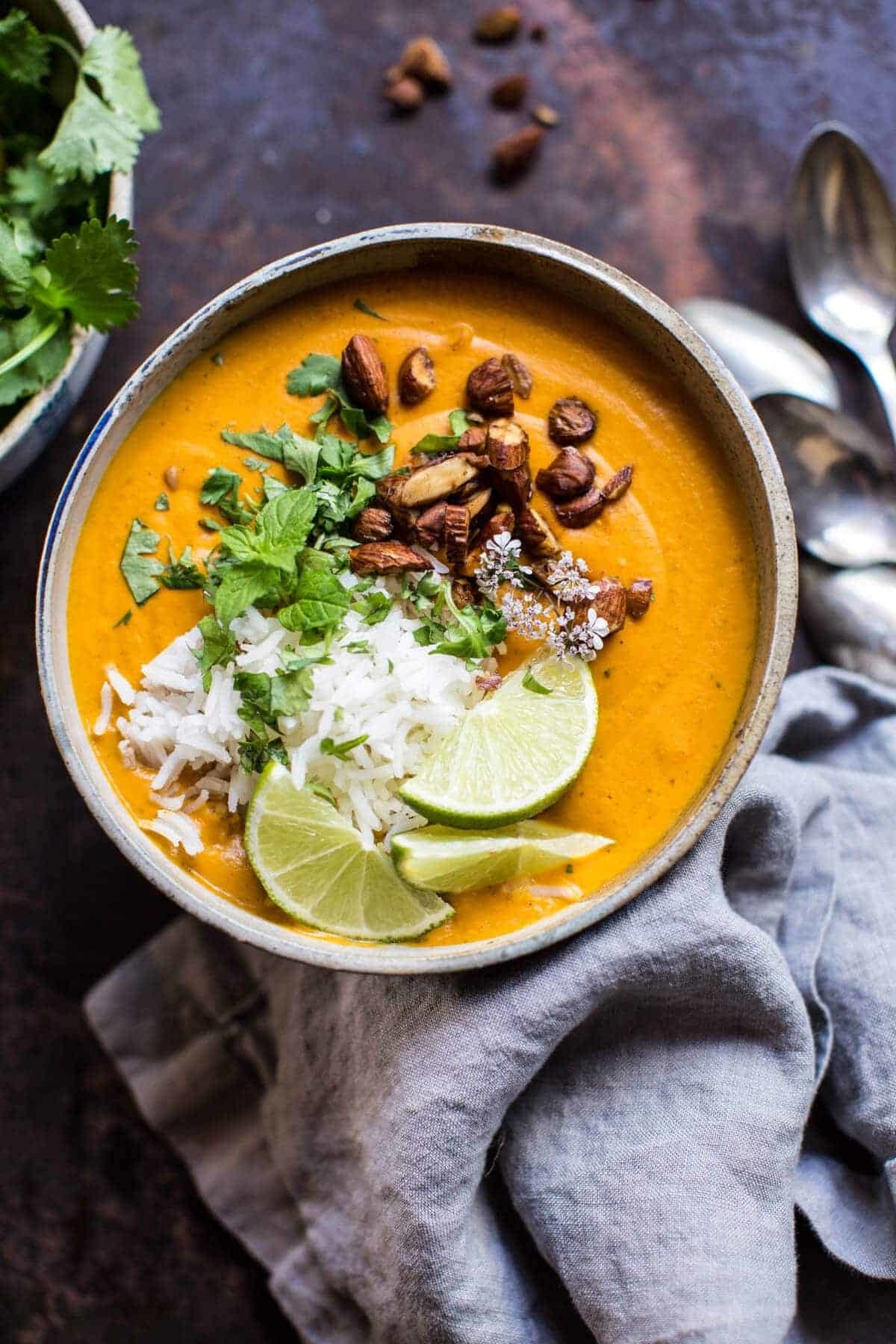 A bowl of creamy soup topped with rice, cilantro, lime wedges, nuts, and seeds on a textured surface with spoons beside it