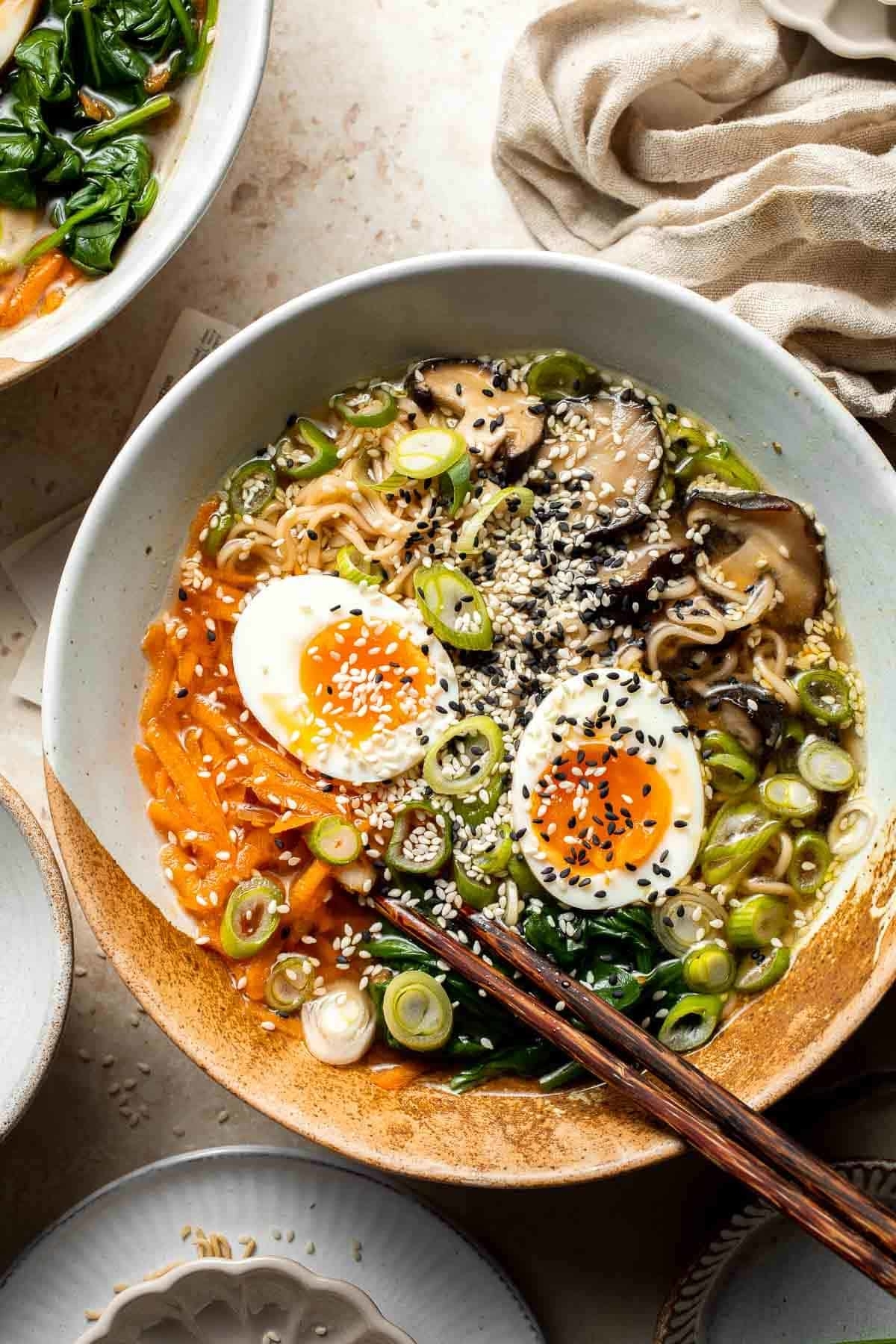 Bowl of ramen with noodles, eggs, mushrooms, scallions, and sesame seeds, garnished with chopsticks on the side