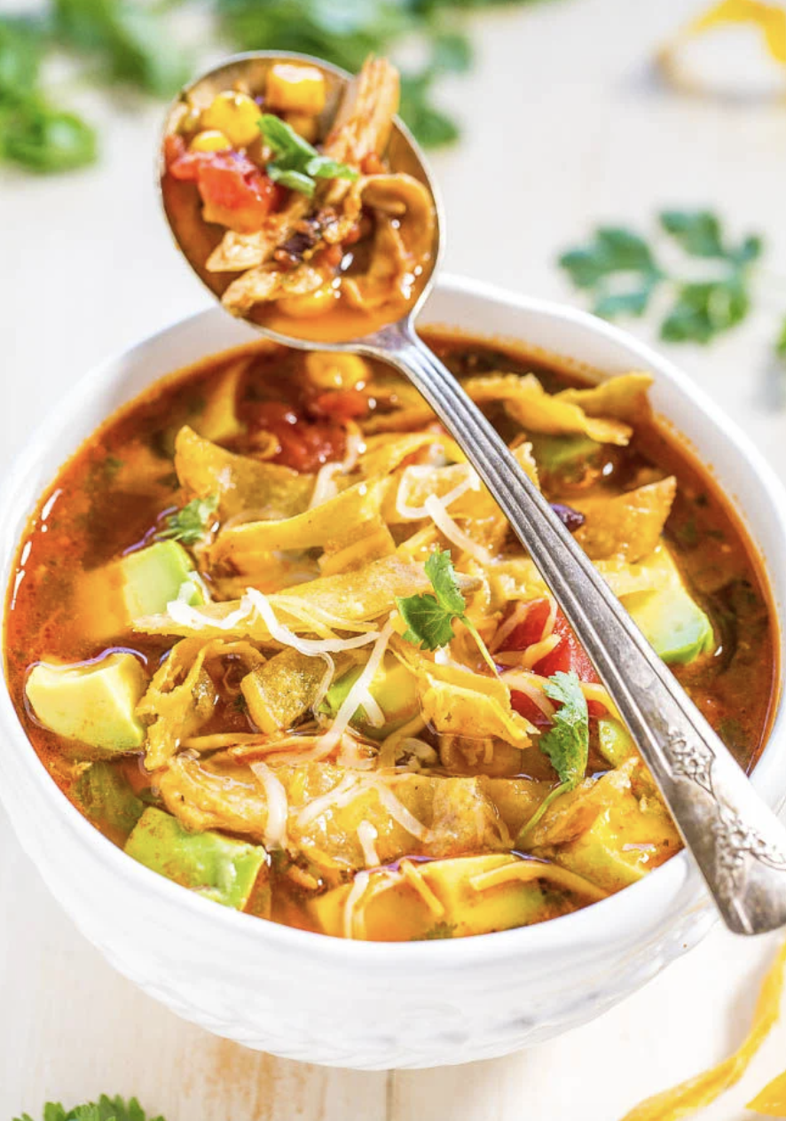 Bowl of chicken tortilla soup with chunks of avocado, shredded cheese, and cilantro, with a spoonful lifted above the bowl