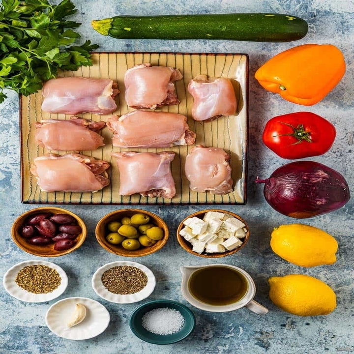 Ingredients for a Mediterranean dish: raw chicken, vegetables, olives, feta cheese, lemons, spices, garlic, and olive oil arranged on a table