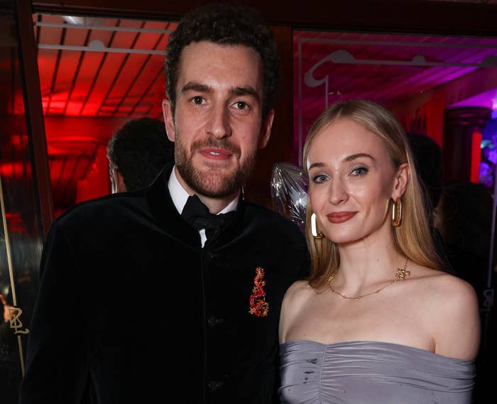 Two people posing at an event; the woman wears a stylish off-shoulder gown, the man in a velvet jacket with a brooch. Both holding drinks