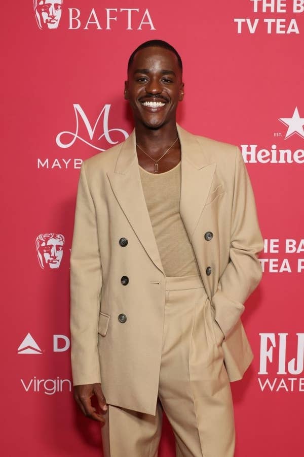 Ncuti smiling on a red carpet wearing a stylish suit, one hand in a pocket