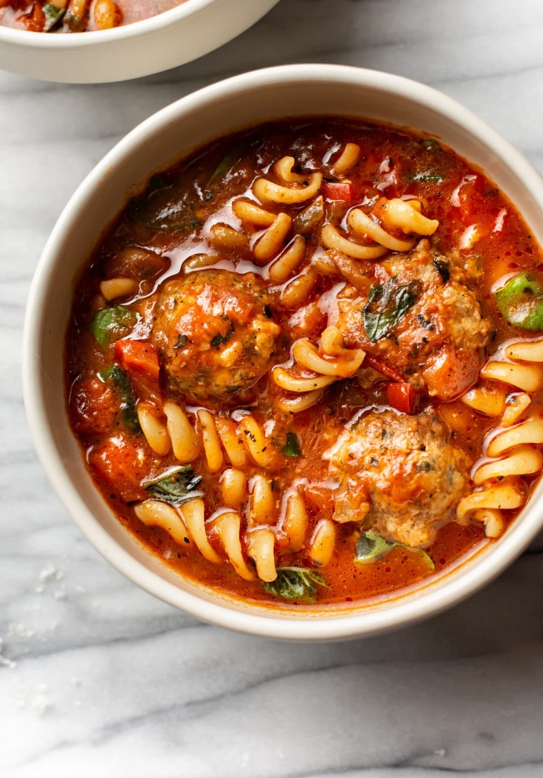 A bowl of pasta soup with rotini, meatballs, and vegetables in a tomato-based broth on a marble surface