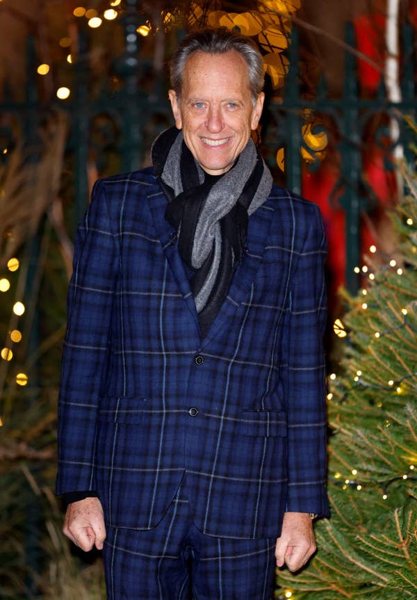 Richard in a stylish plaid suit and scarf, smiling, at an event with decorative lights and foliage in the background