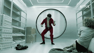 Person in red outfit energetically dances in futuristic room, while another person observes from floor